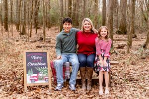 Family of three smiling together in a wooded setting with a Merry Christmas chalkboard sign during a holiday photo session.
