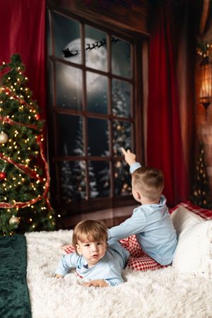 A child in light blue pajamas lies on white fur beside a decorated Christmas tree with red curtains in the background.