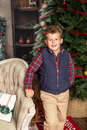 Holiday portrait by decorated Christmas tree with plaid shirt, navy vest and khaki pants, standing next to armchair.