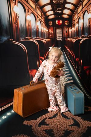 Toddler girl in pajamas holding a teddy bear and vintage suitcase in a decorated train corridor backdrop.