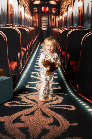Toddler in pajamas holding a stuffed animal stands in a decorated train car aisle with red seats and ornate carpet.