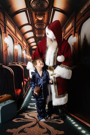 Child in pajamas meets Santa Claus in a festive train corridor decorated for Christmas.