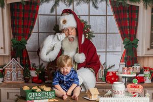Toddler in blue pajamas sits with Santa Claus amid Christmas cookies and festive holiday decorations.