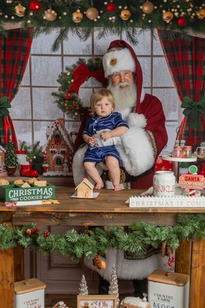 Toddler sitting on Santa Claus lap surrounded by Christmas cookies and festive holiday decorations.