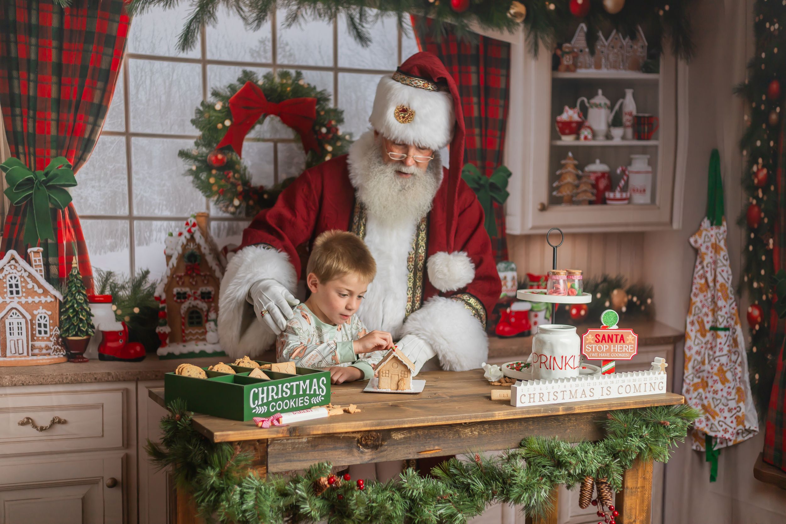 A young boy sits with Santa Claus at a festive wooden table decorating Christmas cookies in a holiday-themed kitchen.