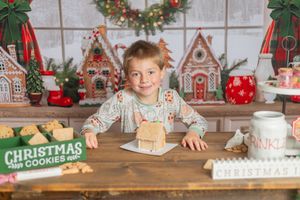 Young child smiles at Christmas cookie decorating table with gingerbread houses and festive holiday backdrop.