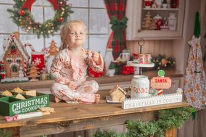 Toddler in Christmas pajamas sits on table with holiday decorations, gingerbread house, and Santa sign in festive kitchen.