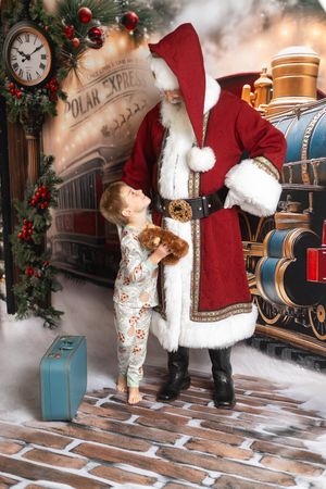 A young child in pajamas holding a teddy bear looks up at Santa Claus beside a Polar Express train backdrop.