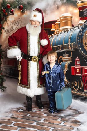 Young boy in blue pajamas with suitcase poses with Santa Claus in front of a Polar Express train backdrop.