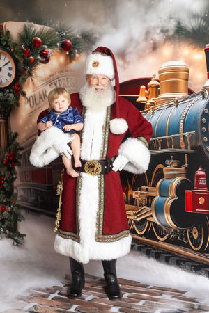 Santa Claus holding a baby in front of a Polar Express train backdrop with festive Christmas decorations.