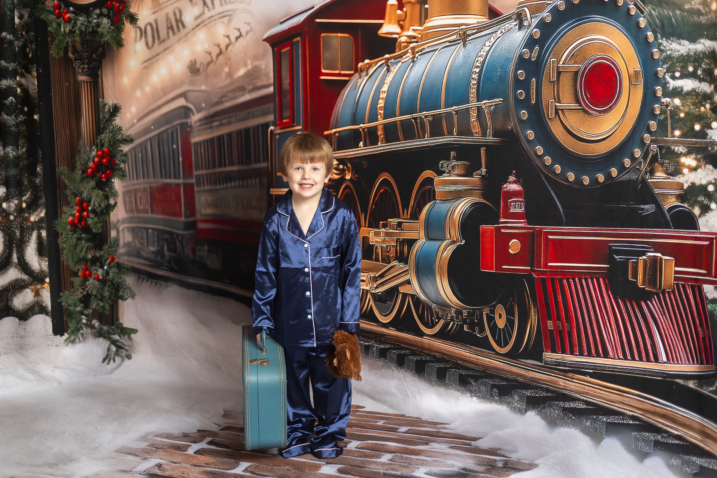 Boy in blue pajamas holding a suitcase beside a Polar Express train mural backdrop.