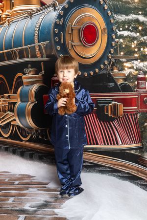 A young child in blue pajamas holds a teddy bear beside a festive Christmas train display with snow.