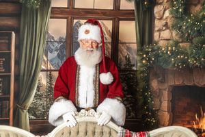 Santa Claus standing behind a chair in a festive room with a fireplace and Christmas tree.