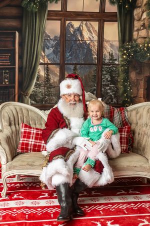 A cheerful toddler sits on Santa Claus's lap on a festive couch with Christmas decorations and snowy mountain backdrop.