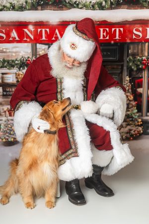 Santa Claus crouching beside a golden retriever dog in a festive Christmas holiday photo setting.