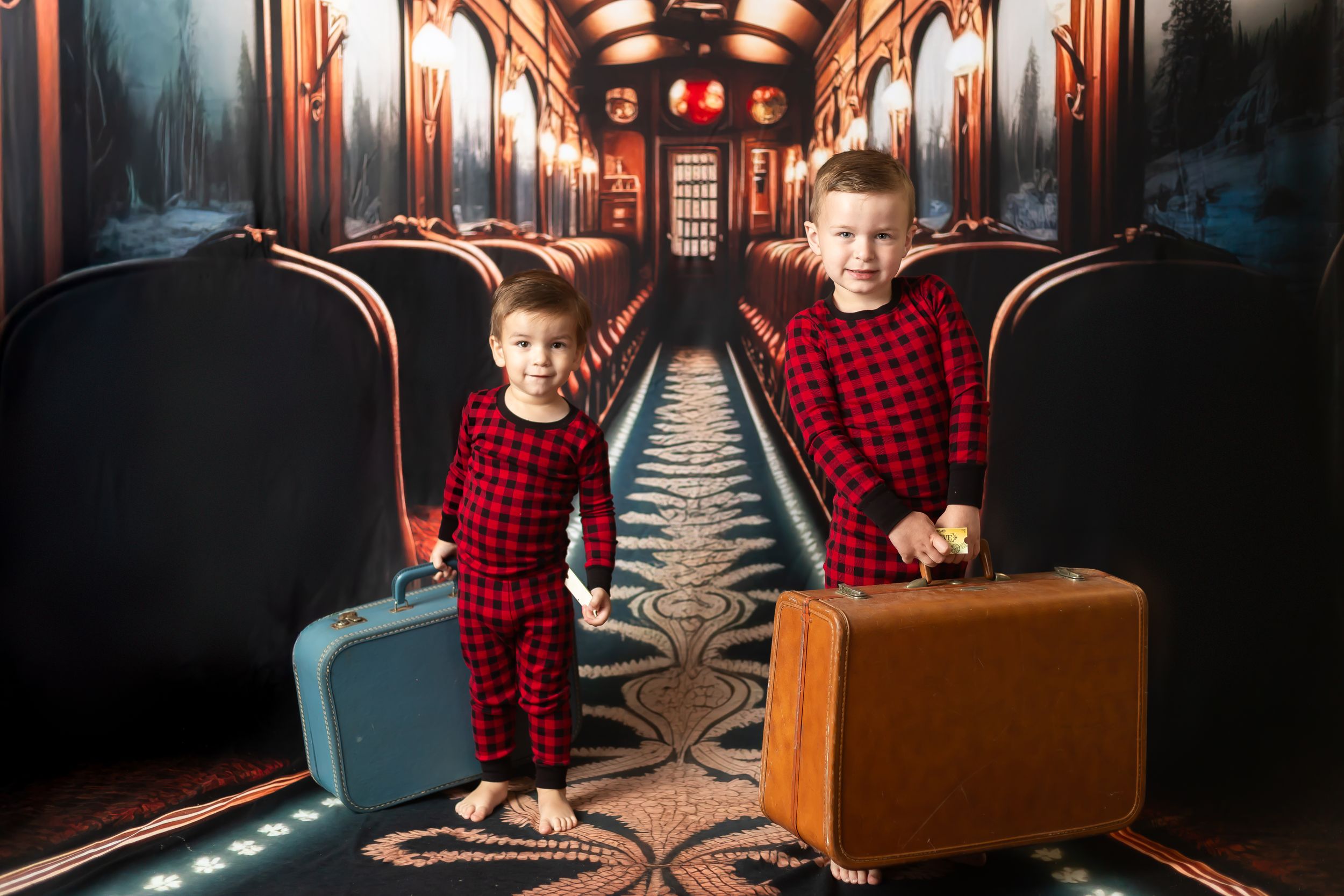 Two young boys in matching red plaid pajamas holding vintage suitcases in front of a festive train corridor backdrop.