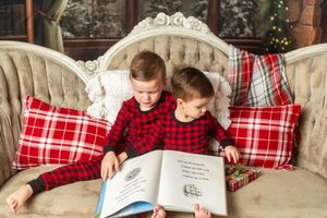 Two young boys in matching red plaid pajamas reading a book together on a festive holiday couch.