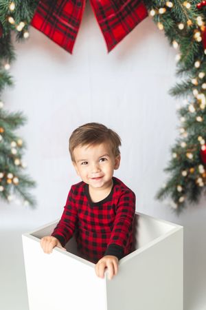 Smiling toddler in red plaid pajamas sitting in a white box surrounded by festive Christmas garland and decorations.