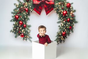 Toddler in red plaid shirt sitting in white box with festive Christmas wreath and red ornaments backdrop.