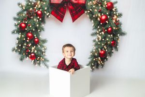 Toddler boy in red plaid outfit sitting in white box with large Christmas wreath and red ornaments behind him.