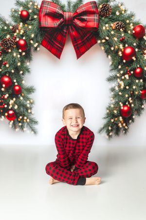 A smiling boy in red plaid pajamas sits cross-legged in front of a large Christmas wreath with red ornaments and bow.