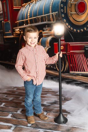 A smiling young boy in a plaid shirt and jeans stands beside a lamp post with a vintage steam train backdrop.