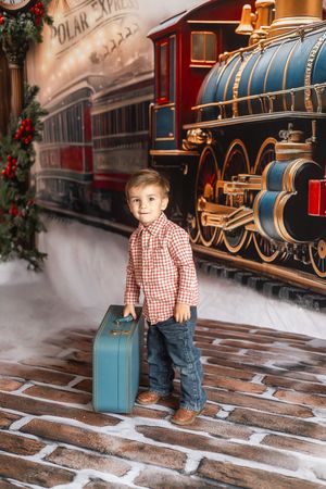 Young boy holding a blue suitcase in front of a Polar Express train backdrop with Christmas decorations.