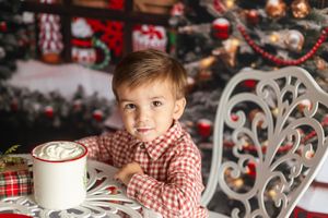 Young boy in red plaid shirt sits at white table with hot cocoa near decorated Christmas tree.