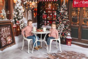 Two children sit at a white table in a festive Christmas setting with decorated trees and a Hot Cocoa sign.