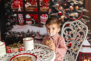 Young boy in red plaid shirt eating cookies near Christmas tree with holiday decorations and gifts.