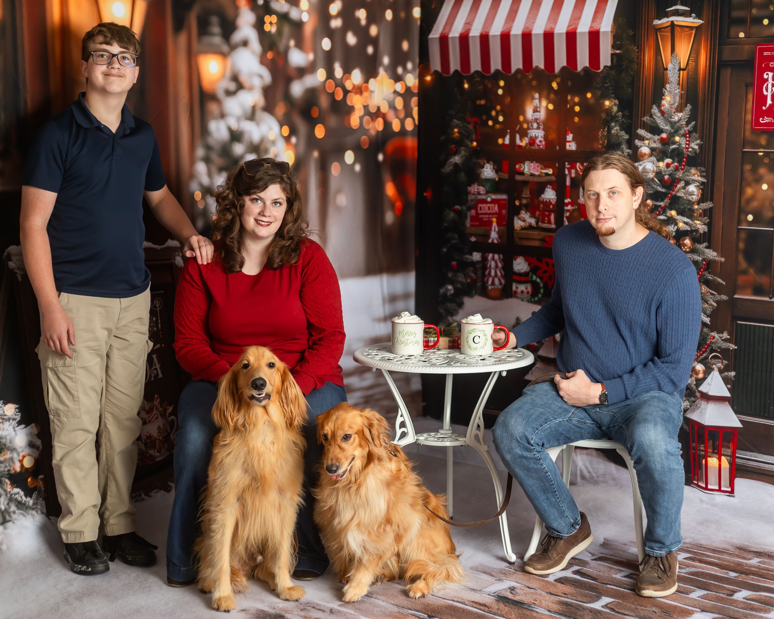 Family of three poses with two golden retrievers in a festive Christmas studio portrait with holiday decorations.