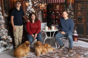 Family of three poses with two golden retrievers in a festive Christmas photo setting with decorated trees.