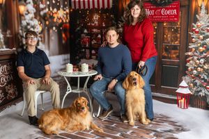 Family of three with two golden retrievers posing in a festive Christmas photo studio with holiday decor.