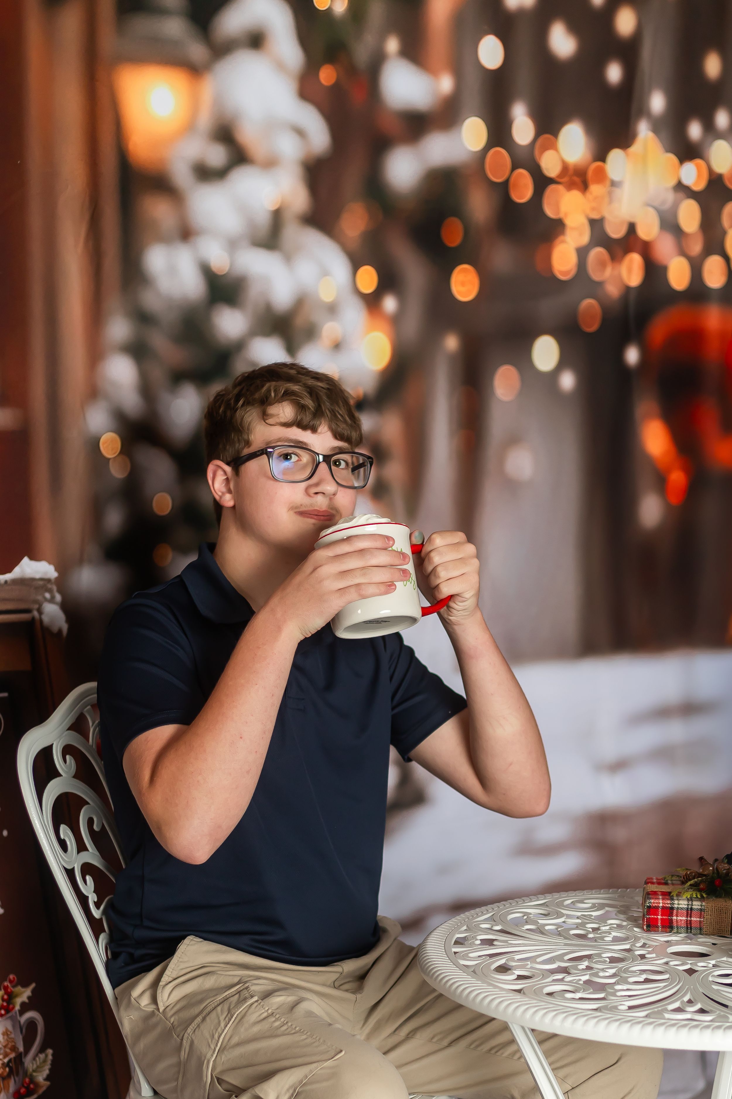 Teen boy in glasses sipping a holiday drink at a white table with Christmas lights and snowy decor in background.