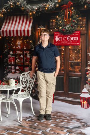 Teen boy in glasses and khaki pants poses by a festive Hot Cocoa shop with Christmas decor and twinkling lights.