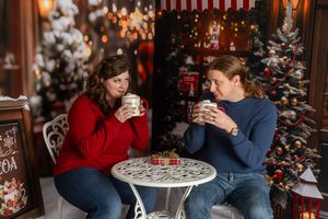 Two people in festive sweaters enjoy hot cocoa at a white table surrounded by decorated Christmas trees indoors.