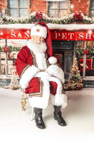 Santa Claus sits in a chair holding a bell outside a festive Christmas pet shop decorated with snow and holiday lights.
