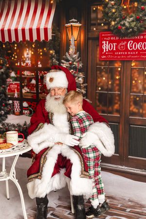 Child in plaid pajamas sitting on Santa Claus lap in a festive Christmas shop with hot cocoa sign.