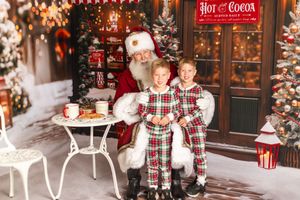 Two children in plaid pajamas sit with Santa Claus in a festive Christmas setting with trees and hot cocoa sign.