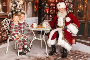 Two children in plaid pajamas sit with Santa Claus at a festive table with cookies and hot cocoa in a Christmas setting.
