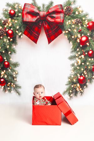 Adorable baby sitting inside a red gift box beneath a festive Christmas wreath with red ornaments and plaid bow.