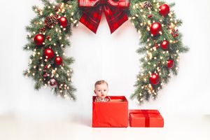 Baby sitting inside a red gift box under a festive Christmas wreath with red ornaments and lights.