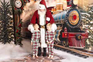 Two children in plaid pajamas pose with Santa Claus in front of a festive Polar Express train backdrop.
