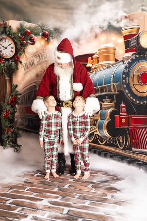 Two children in plaid pajamas pose with Santa Claus in front of a festive Polar Express train backdrop.