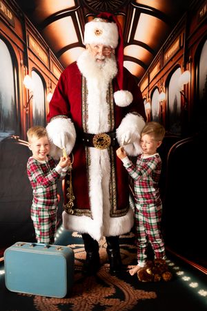 Two children in plaid pajamas pose with Santa Claus in a festive train car setting with a blue suitcase.
