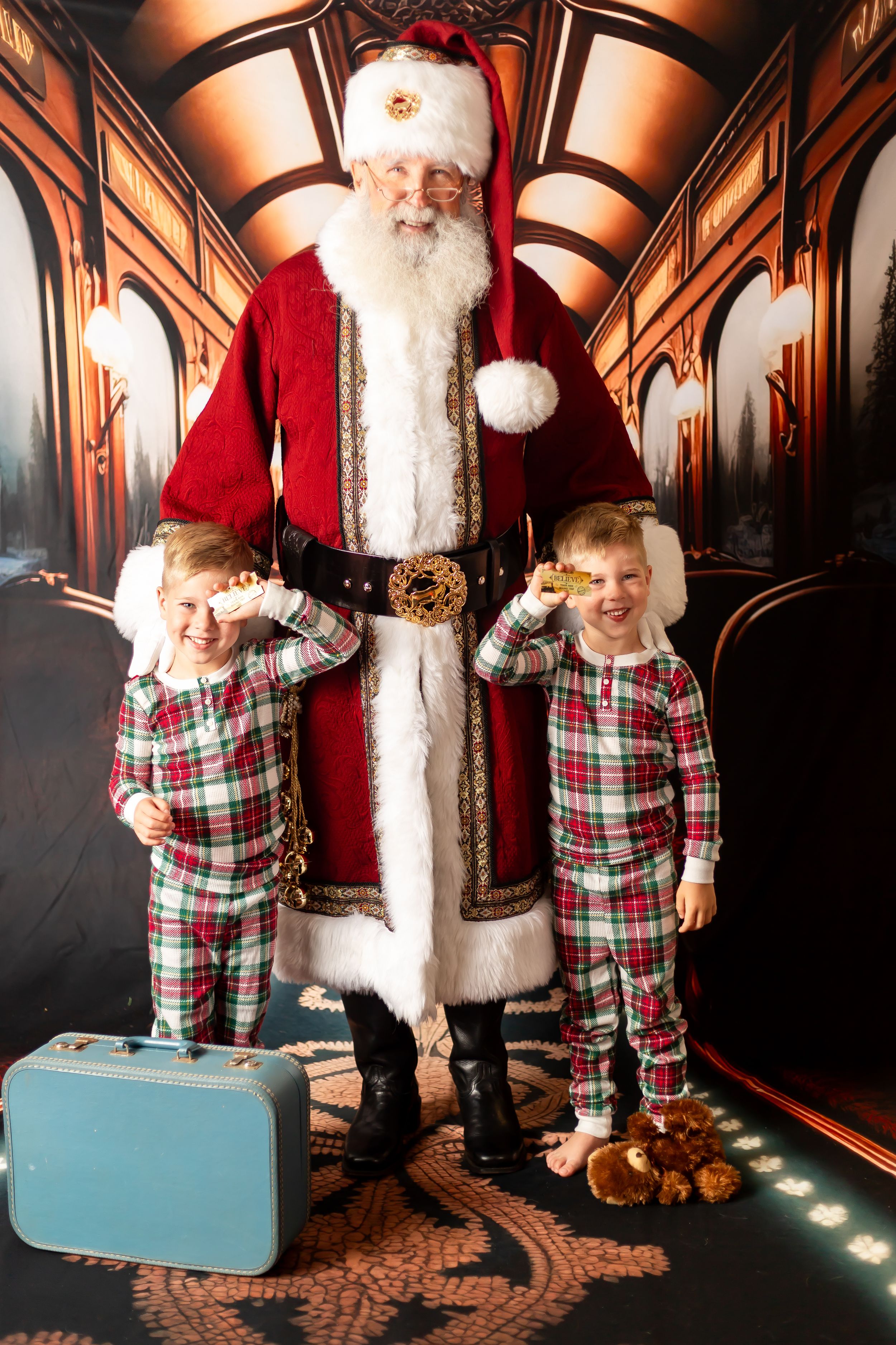 Two children in plaid pajamas pose with Santa Claus, one holding a golden ticket, beside a blue suitcase.
