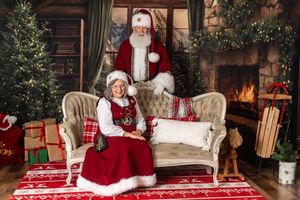 Santa Claus and Mrs. Claus pose on a vintage sofa by a fireplace with Christmas trees and gifts.