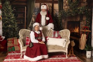Santa Claus and Mrs. Claus pose together in a festive holiday setting with Christmas trees and a fireplace.