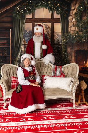 Santa and Mrs. Claus pose together in a festive holiday setting with Christmas decorations, fireplace, and red reindeer rug.