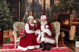 Santa and Mrs. Claus sit together on a vintage sofa surrounded by Christmas trees, gifts, and a warm fireplace.